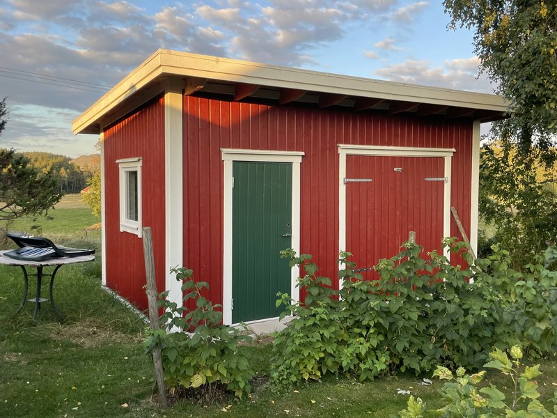 Rödmålad redskapsbod med vita knutar, grön dörr och pulpettak, i kvällsljus